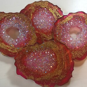 May include: Four red, white, and gold glitter resin coasters with a ring in the center. The coasters are shaped like agate slices.