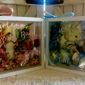 May include: Two white shadow boxes with glass fronts, each containing a floral arrangement and a small silver spoon. The box on the left has a pink ribbon and the text "welcome Baby" in pink script. The box on the right has a blue ribbon and the text "welcome to my crib" in blue script.