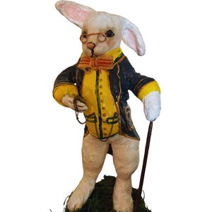 May include: A white rabbit figurine wearing a dark blue coat with yellow trim, a yellow vest, and a striped bow tie. The rabbit has wire-rimmed glasses, holds a thin brown cane, and carries a pocket watch on a chain, standing on a green base.