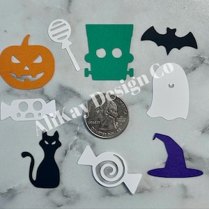 May include: A collection of Halloween-themed felt cutouts, including an orange jack-o'-lantern, a green Frankenstein's monster, a black bat, a white ghost, a black cat, and various candies. The image also includes a quarter for size reference. The text "Alikay Design Co" is visible.