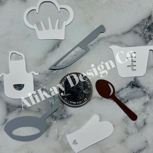 May include: A set of white paper cutouts of kitchen items, including a chef's hat, apron, knife, measuring cup, and oven mitt. The items are arranged on a gray surface with a US quarter for scale.