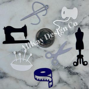 May include: A set of nine white and black die-cut paper shapes for crafting. The shapes include a sewing machine, a spool of thread, a needle and thread, a pair of scissors, a pincushion, a measuring tape, and a dress form.