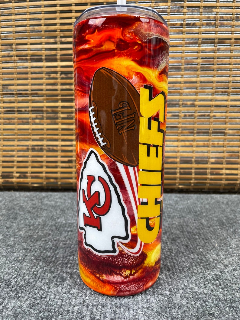May include: A red, yellow and orange swirl design tumbler with a Chiefs logo and a football. The tumbler has a clear lid and a straw.