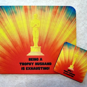 May include: A yellow and red tie-dye design with a gold statue of a person holding an award. The text "Being a Trophy Husband is Exhausting!" is printed below the statue.
