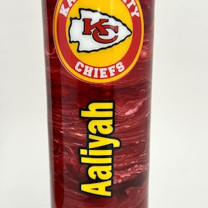 May include: A red and white marbled tumbler with a Kansas City Chiefs logo and the name "Aaliyah" printed on it. The tumbler has a lid with a hole for a straw.
