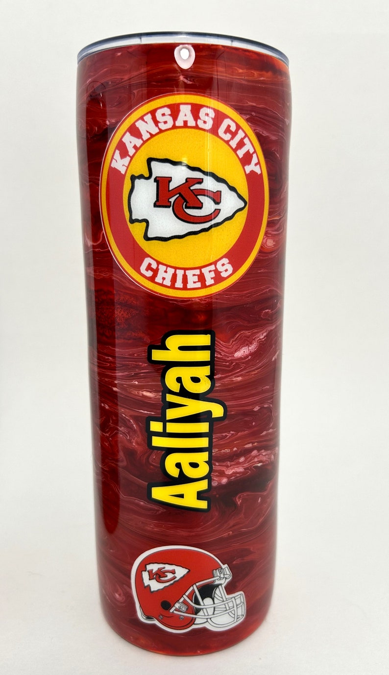 May include: A red and white marbled tumbler with a Kansas City Chiefs logo and the name "Aaliyah" printed on it. The tumbler has a lid with a hole for a straw.