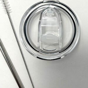 May include: A clear plastic lid with a sliding closure for a reusable tumbler. The lid is shown with a stainless steel straw and a cleaning brush.