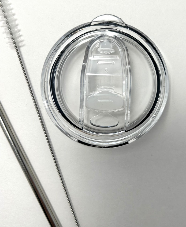 May include: A clear plastic lid with a sliding closure for a reusable tumbler. The lid is shown with a stainless steel straw and a cleaning brush.
