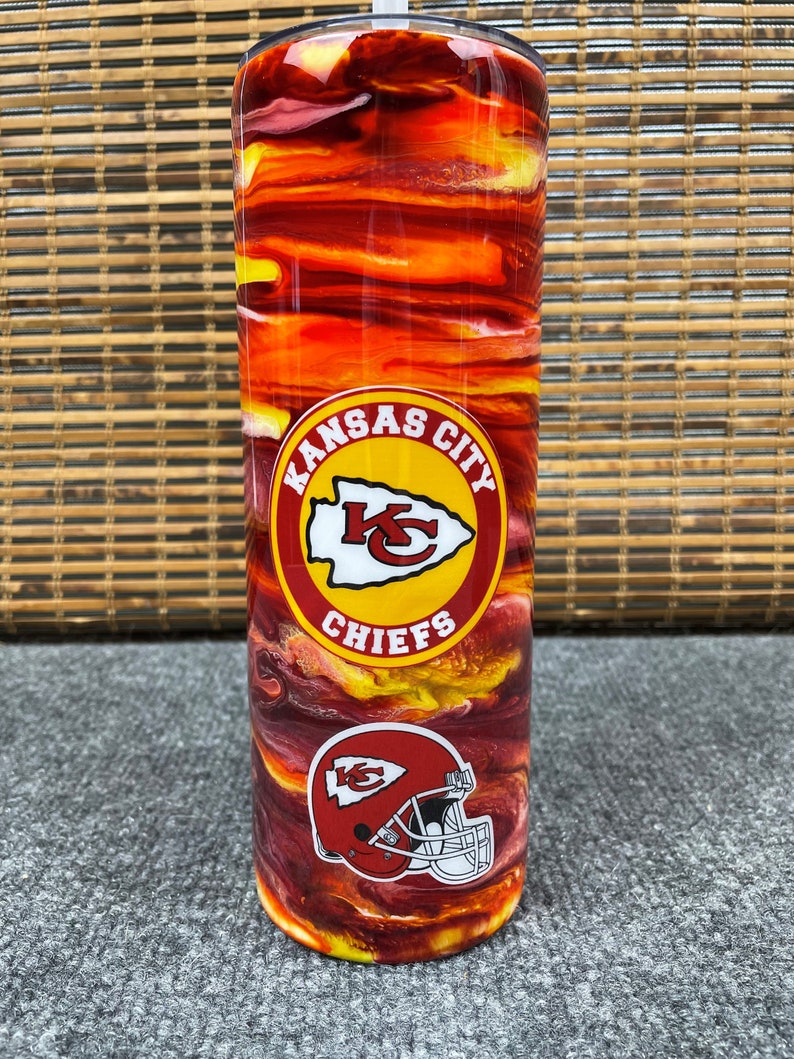 May include: A red and yellow stainless steel tumbler with a Kansas City Chiefs logo and a football helmet. The tumbler has a swirl pattern.