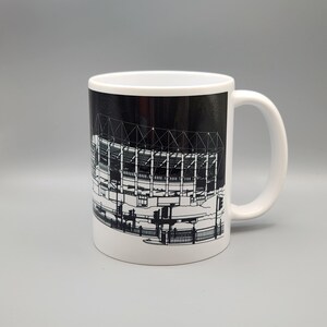 May include: White ceramic mug with a black and white illustration of a stadium on the front. The stadium is outlined in white and the background is black.