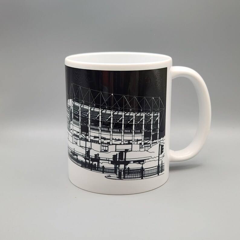 May include: White ceramic mug with a black and white illustration of a stadium on the front. The stadium is outlined in white and the background is black.