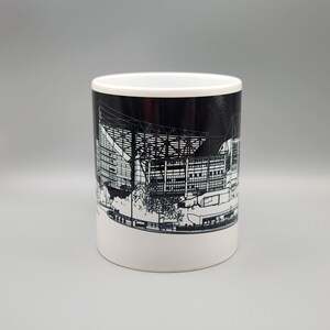 May include: A white ceramic mug with a black and white illustration of a stadium on the side. The stadium is outlined in white and the background is black.