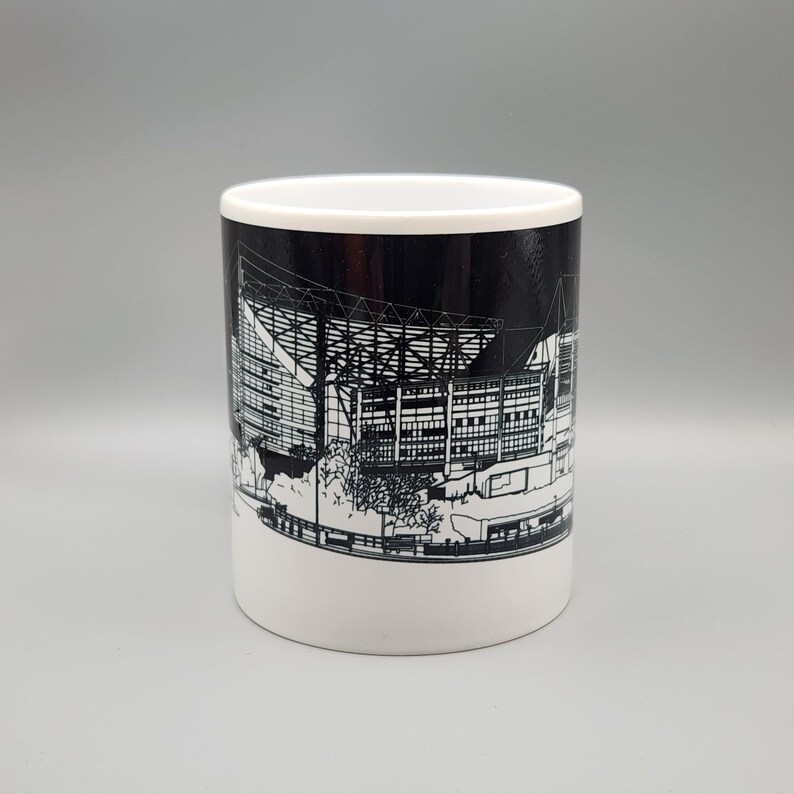 May include: A white ceramic mug with a black and white illustration of a stadium on the side. The stadium is outlined in white and the background is black.