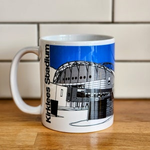 Könnte beinhalten: Weiße Keramiktasse mit einer blauen und schwarzen Illustration eines Stadions mit dem Text "Kirklees Stadium" an der Seite.