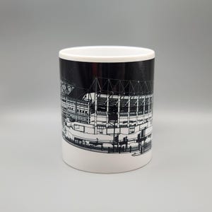 May include: A white ceramic mug with a black and white illustration of a stadium on the side. The stadium is outlined in white and the background is black.