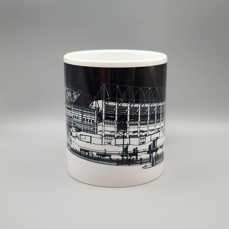 May include: A white ceramic mug with a black and white illustration of a stadium on the side. The stadium is outlined in white and the background is black.