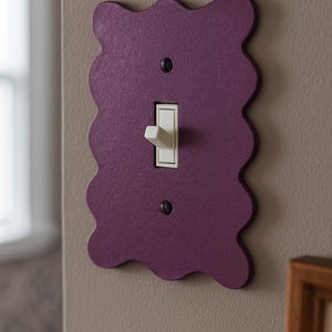 May include: A decorative purple light switch cover with a wavy edge. The cover has a standard light switch and is mounted on a neutral-colored wall. The switch is off.