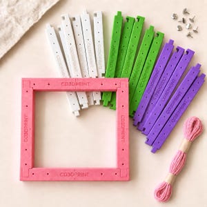 May include: A square frame kit with a pink base, featuring white, green, and purple interlocking plastic bars labeled CO3DPRINT. The set includes small metal pins and a bundle of pink embroidery thread for assembly and crafting projects.