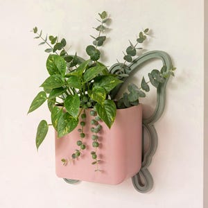 May include: A pink wall-mounted planter with a decorative gray frame, filled with green plants. The planter holds a variety of greenery, including trailing string of pearls and leafy plants. The design is modern and adds a touch of nature to interior decor.