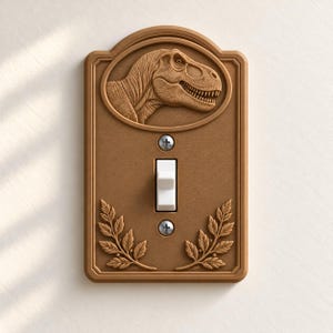 May include: A rectangular light switch cover plate in a matte brown finish. The top features a relief carving of a Tyrannosaurus Rex head inside an oval frame. The bottom displays two symmetrical leafy branches. A white toggle switch is centered.