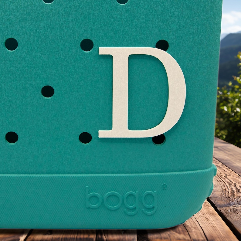 May include: Teal blue tote bag with a white letter D on the front. The bag has a textured surface with small holes. The bottom of the bag has the word "bogg" printed in white.
