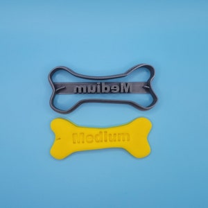 May include: A gray plastic dog bone cookie cutter with the word "Medium" embossed on it. Next to it is a yellow plastic dog bone with the word "Medium" embossed on it.