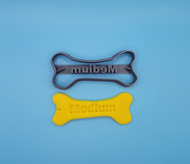 May include: A gray plastic dog bone cookie cutter with the word "Medium" embossed on it. Next to it is a yellow plastic dog bone with the word "Medium" embossed on it.