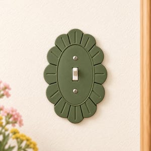 May include: A matte olive green light switch cover plate featuring a scalloped, flower-like edge design. The oval-shaped plate is secured with two silver-toned metal screws and includes a single white toggle switch in the center.