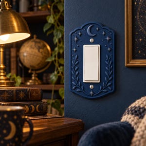 May include: A dark blue rectangular light switch cover plate featuring a raised relief design of a crescent moon, various stars, and leafy vine branches. The plate is secured with two silver-toned screws and surrounds a white rocker switch.