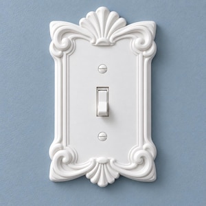 May include: A white rectangular light switch wall plate featuring an ornate, raised border with scrollwork and shell-like motifs at the top and bottom. The plate includes a central toggle switch and two matching white screws for installation.