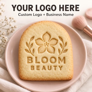 May include: A light-colored, arch-shaped cookie featuring an embossed design of a central flower flanked by two leafy sprigs, with the text BLOOM BEAUTY debossed below. The cookie is centered on a smooth, pale pink plate.