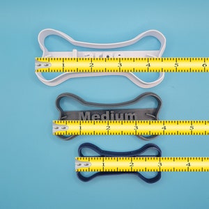 May include: Three dog bone cookie cutters in different sizes: large, medium, and small. The cookie cutters are made of white, gray, and blue plastic. Each cookie cutter is measured with a yellow ruler.