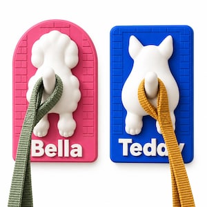 May include: Two wall-mounted hooks featuring white dog rear ends on rectangular bases. The left hook is pink with the name Bella and holds a green leash. The right hook is blue with the name Teddy and holds a yellow leash. Both have brick patterns.