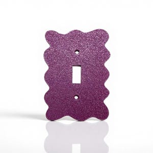 May include: A decorative purple glitter-covered light switch cover with a wavy edge design. The cover has a rectangular opening for the switch and two screw holes. The switch plate is shown against a white background.