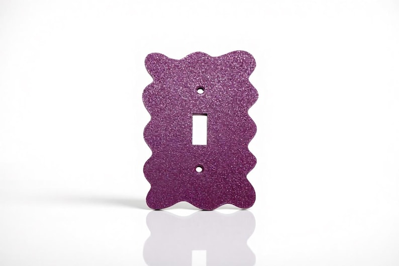 May include: A decorative purple glitter-covered light switch cover with a wavy edge design. The cover has a rectangular opening for the switch and two screw holes. The switch plate is shown against a white background.