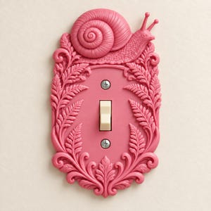 May include: A pink light switch plate featuring a raised snail figure at the top and intricate fern leaf patterns along the border. The rectangular plate includes a central cream-colored toggle switch secured by two visible metal screws.