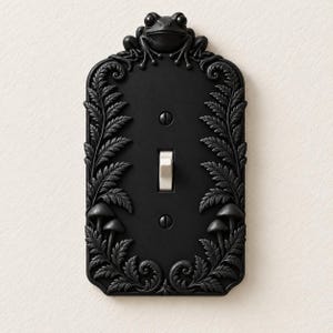 May include: A matte black light switch plate featuring a sculpted frog at the top, surrounded by embossed fern fronds and small mushrooms along the sides. The rectangular cover includes a central toggle switch and two mounting screws.
