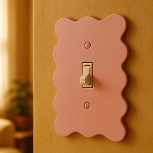 May include: A decorative light switch cover in a soft pink hue with a wavy edge design. The switch and screws are a cream color, contrasting with the pink. The cover is mounted on a mustard-yellow wall.
