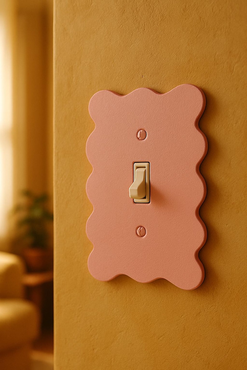 May include: A decorative light switch cover in a soft pink hue with a wavy edge design. The switch and screws are a cream color, contrasting with the pink. The cover is mounted on a mustard-yellow wall.