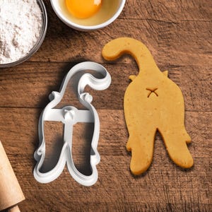 May include: A white plastic cookie cutter in the shape of a cat's rear end, next to a baked cookie of the same shape. The cookie has an 'X' mark. Ingredients like flour and an egg are in the background.