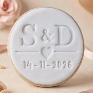 May include: A round sugar cookie topped with smooth white icing. The center features the embossed letters S & D above a small heart symbol and a horizontal line. Below the line, the date 14-11-2026 is embossed in a clean, sans-serif font.