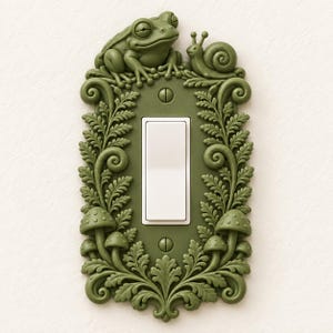 May include: An olive green light switch cover plate featuring a raised relief design of a frog and a snail at the top, surrounded by detailed fern fronds and small mushrooms along the sides and bottom, secured with two matching screws.