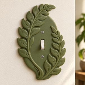 May include: An olive green light switch cover plate featuring a raised, embossed fern leaf design that frames the central toggle switch. The plate is secured with two silver-colored metal screws positioned above and below the white toggle switch.