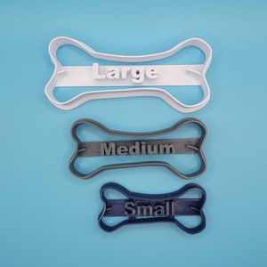May include: Three dog bone-shaped cookie cutters, one white, one gray, and one blue. The white cutter is labeled "Large", the gray cutter is labeled "Medium", and the blue cutter is labeled "Small".
