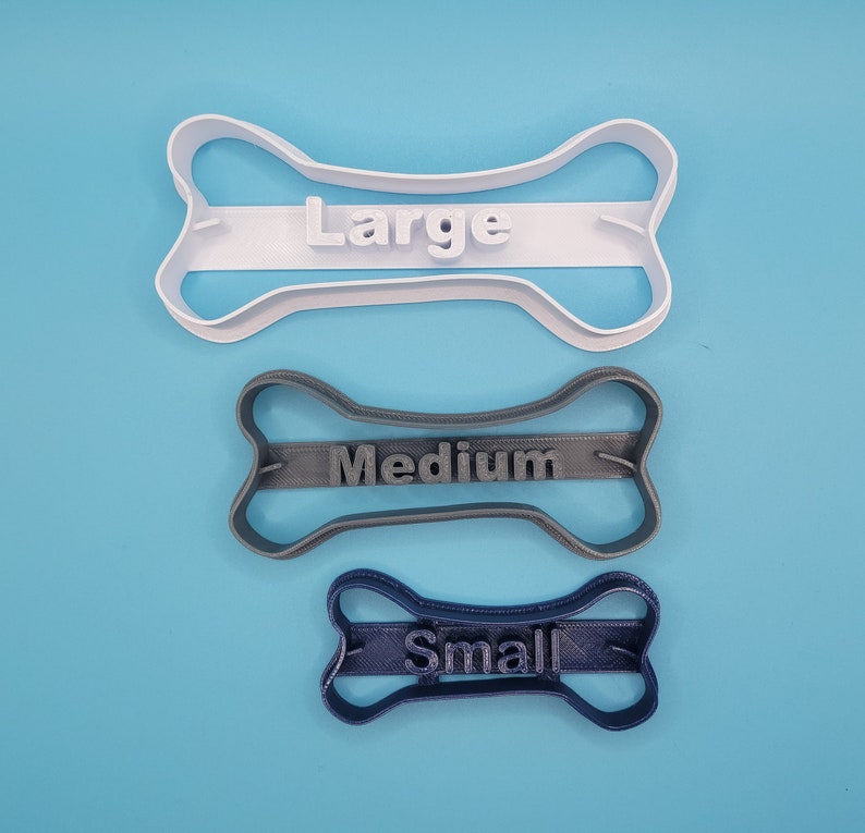 May include: Three dog bone-shaped cookie cutters, one white, one gray, and one blue. The white cutter is labeled "Large", the gray cutter is labeled "Medium", and the blue cutter is labeled "Small".