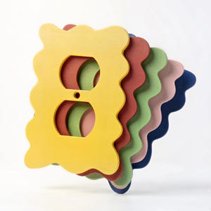 May include: A stack of colorful, wavy-edged outlet covers in shades of yellow, red, green, pink, and blue. Each cover has two circular openings and a small hole for the screw.