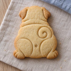 May include: A light tan, baked sugar cookie shaped like the rear view of a pug dog. The cookie features embossed details including the dog's rounded back, a curled tail, hind legs, and a small asterisk-shaped mark centered on the lower portion.