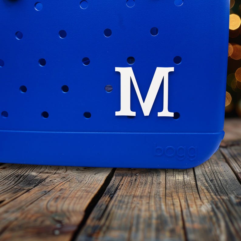 May include: A bright blue tote bag with a white letter "M" on the front. The bag has a textured surface with small holes. The bag is sitting on a wooden surface.