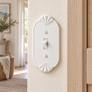 May include: A white oval light switch wall plate featuring a raised shell-like fan design at the top and bottom. The plate is secured with two flat-head screws and surrounds a single white toggle switch, mounted on a light-colored wall surface.