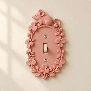 May include: A dusty pink light switch cover plate featuring a sculpted mouse perched at the top and a decorative border of clover leaves and stems. The plate includes a central toggle switch and two silver-colored mounting screws.